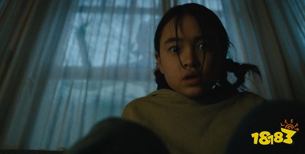Ruth, a young girl, stares directly into the camera as a shadowy figure leans against the window.