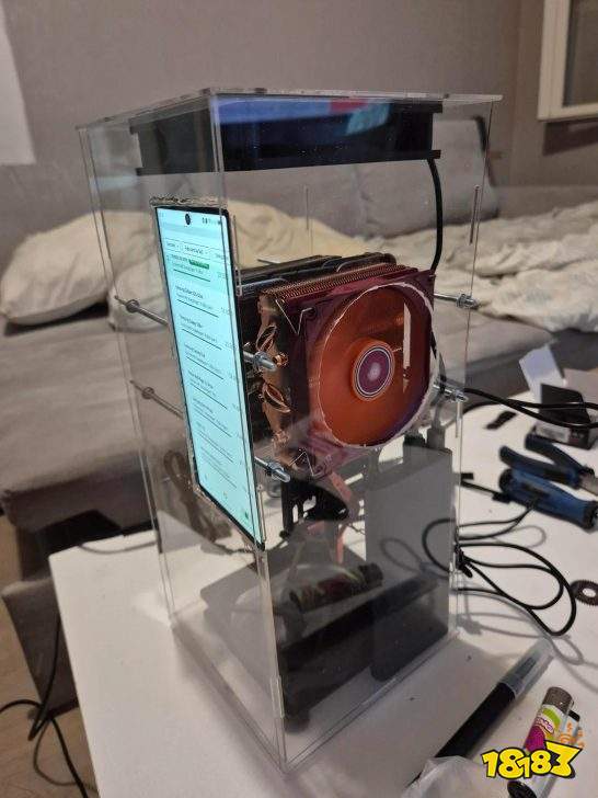A smartphone is used as a display in a homemade computer encased in a transparent container, featuring a prominent red CPU cooler.