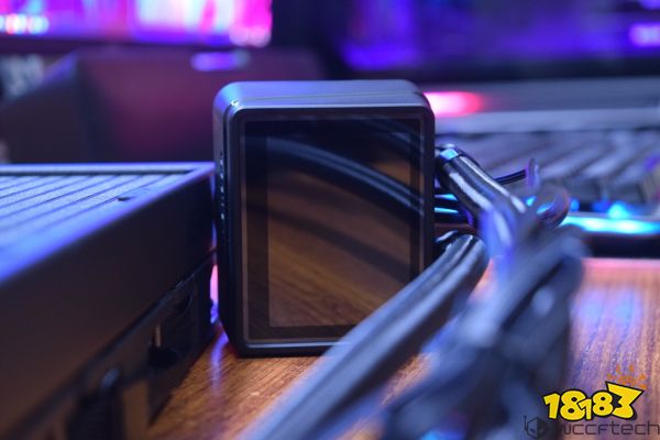 A close-up of a black, square display unit on a computer cooling system with visible braided cables in a tech setup.