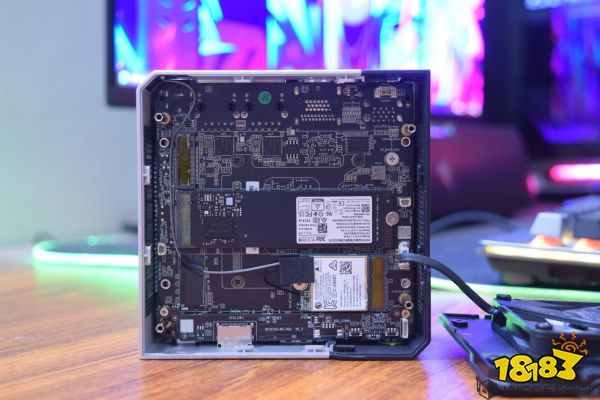 The interior of an unbranded mini PC case showing the motherboard with a Kingston M.2 NVMe SSD and a Wi-Fi card installed on a wooden desk, with blurred RGB-lit screens in the background.