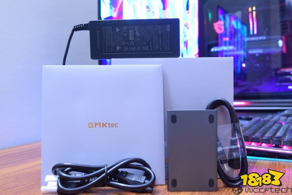 A GMKtec product box with branding, alongside a power adapter, cables, and a metallic peripheral, is displayed on a wooden surface with a colorful PC setup in the background.
