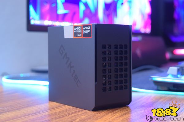A GMKtec mini PC with AMD Ryzen 9 HX and Radeon graphics logos is displayed on a wooden desk with a colorful monitor in the background.