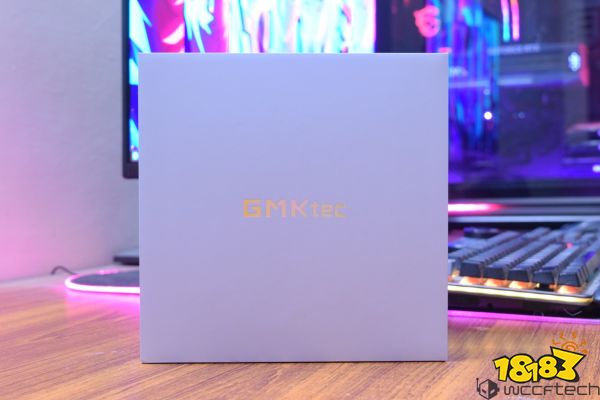 A GMKtec product box is placed on a wooden desk in front of a gaming setup with a colorful, illuminated background.