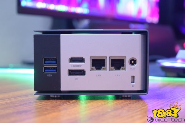 The back panel of a mini PC showing two USB ports, HDMI, DP, two LAN ports, and a DCIN connector on a wooden surface with a blurred colorful background.