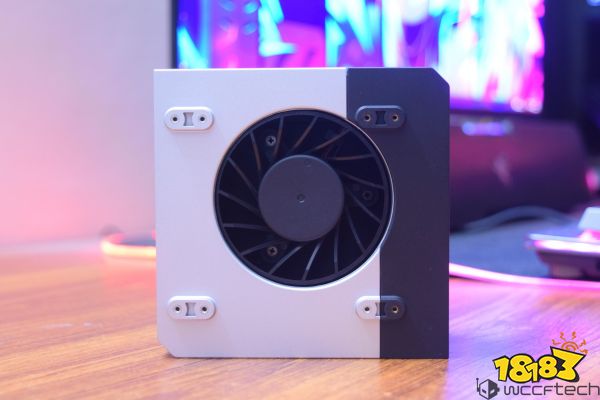 A close-up of a small desktop cooling fan with a circular fan design, set against a two-tone, white and black casing on a wooden surface, with a soft-focus computer setup in the background.